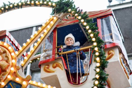 Foto de Feliz linda niña preescolar a caballo en el carrusel de la rueda del ferris en la feria o mercado de Navidad, al aire libre. Pequeño niño divirtiéndose en el mercado tradicional de Navidad familiar en Alemania. - Imagen libre de derechos