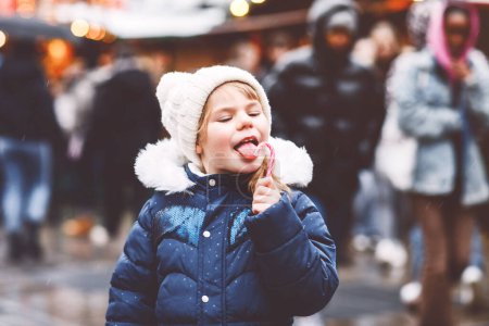 Foto de Niña linda preescolar con bastón de caramelo de un puesto de dulces en el mercado de Navidad. Niño feliz en el mercado familiar tradicional en Alemania. Preescolar en ropa colorida de invierno durante las nevadas. - Imagen libre de derechos