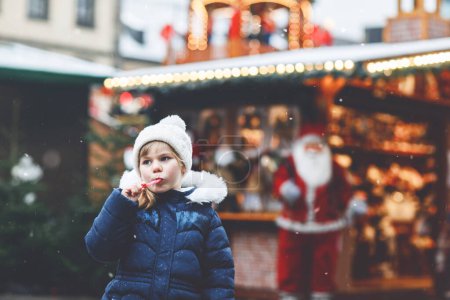 Téléchargez les photos : Petite fille mignonne préscolaire avec canne à bonbons d'un stand de bonbons sur le marché de Noël. Enfant heureux sur le marché familial traditionnel en Allemagne. Préscolaire en vêtements d'hiver colorés pendant les chutes de neige. - en image libre de droit