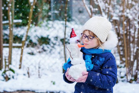 Foto de Cute little preschool girl with glasses making mini snowman. Adorable healthy happy child playing and having fun with snow, outdoors on cold day. Active leisure with children in winter. - Imagen libre de derechos