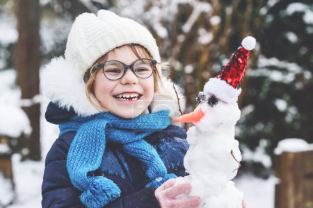Téléchargez les photos : Cute little preschool girl with glasses making mini snowman. Adorable healthy happy child playing and having fun with snow, outdoors on cold day. Active leisure with children in winter. - en image libre de droit