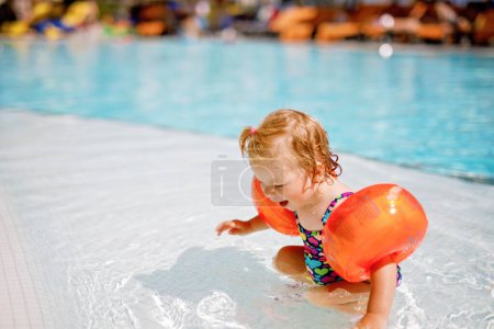 Téléchargez les photos : Petite fille tout-petit avec des nageurs protecteurs jouant dans la piscine extérieure au coucher du soleil. Bébé Enfant apprenant à nager dans la piscine extérieure, éclaboussant d'eau, riant et s'amusant. Vacances en famille - en image libre de droit