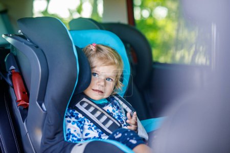 Téléchargez les photos : Adorable petite fille aux yeux bleus assise dans le siège de sécurité de la voiture. Enfant en bas âge en vacances en famille et Jorney. Sourire enfant heureux pendant les embouteillages - en image libre de droit