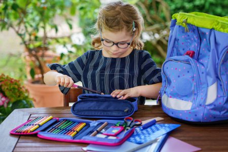 Photo for Happy smiling girl preparing for school her backpack. First day of school. Back to school concept. Little child collecting different supplies like pens and books in a bag or satchel - Royalty Free Image