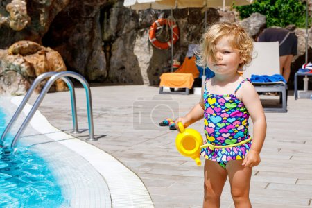 Téléchargez les photos : Jolie petite fille heureuse tout-petit dans la piscine et s'amuser en vacances en famille dans une station hôtelière. Enfant en bonne santé jouant dans l'eau. Bébé en maillot de bain coloré - en image libre de droit