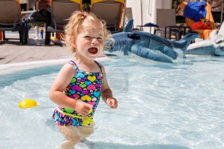 Téléchargez les photos : Petite fille en pleurs bouleversée dans la piscine en vacances en famille dans un hôtel. Triste enfant jouant dans l'eau. Bébé en maillot de bain coloré - en image libre de droit