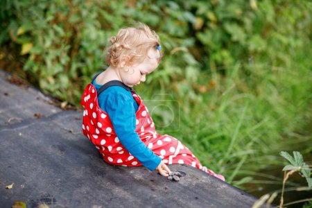 Photo for Cute adorable toddler girl sitting on wooden bridge and throwing small stones into a creek. Funny baby having fun with outdoor games in nature. Active outdoors leisure and activity with little kids - Royalty Free Image