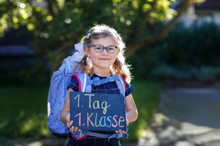 Photo for Happy little kid girl with eye glasses with backpack or satchel and big school bag on the first day of school. Healthy adorable child outdoors. Child holding chalks desk for first schoolday - Royalty Free Image
