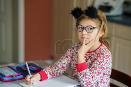 Téléchargez les photos : Petite écolière qui fait ses devoirs à la maison. Joyeux enfant heureux apprenant à écrire, classe primaire. Fille avec des lunettes tenant stylo. Retour à l'école - en image libre de droit