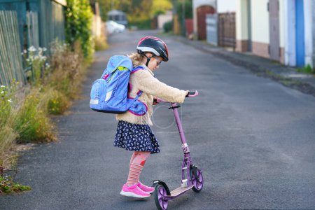 Foto de La niña monta una patineta scooter en su camino de regreso a la escuela. Lindo niño con mochila y con casco de seguridad. Niño de escuela feliz - Imagen libre de derechos