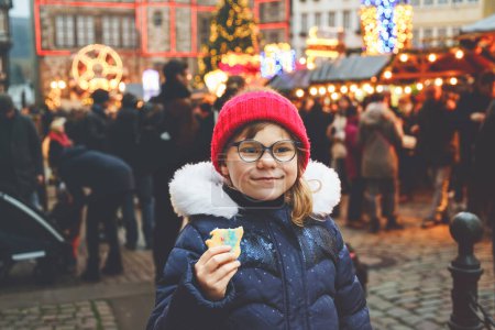 Foto de Niña linda preescolar comiendo galletas de jengibre en el mercado alemán de Navidad. Niño feliz en el mercado familiar tradicional en Alemania, niño riendo en ropa de invierno colorida. - Imagen libre de derechos