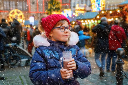 Téléchargez les photos : Petite fille préscolaire mignonne qui boit du punch chaud pour enfants ou du chocolat sur le marché de Noël allemand. Enfant heureux sur le marché familial traditionnel en Allemagne, enfant riant en vêtements d'hiver colorés. - en image libre de droit