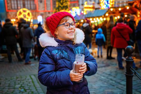 Téléchargez les photos : Petite fille préscolaire mignonne qui boit du punch chaud pour enfants ou du chocolat sur le marché de Noël allemand. Enfant heureux sur le marché familial traditionnel en Allemagne, enfant riant en vêtements d'hiver colorés. - en image libre de droit
