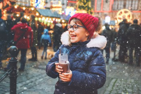 Photo for Little cute preschool girl drinking hot children punch or chocolate on German Christmas market. Happy child on traditional family market in Germany, Laughing kid in colorful winter clothes. - Royalty Free Image