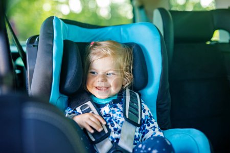 Téléchargez les photos : Adorable petite fille aux yeux bleus assise dans le siège de sécurité de la voiture. Enfant en bas âge en vacances en famille et Jorney. Sourire enfant heureux pendant les embouteillages - en image libre de droit