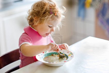 Téléchargez les photos : Adorable petite fille mangeant de la soupe de nouilles aux légumes cuillère. nourriture, enfant, alimentation et concept de développement. Enfant tout-petit mignon, fille avec cuillère assise en chaise haute et apprenant à manger par lui-même. - en image libre de droit