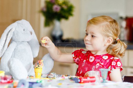 Photo for Adorable cute little toddler girl with colorful clay. Healthy baby playing and creating toys from play dough. Small happy funny child molding modeling clay, making ice cream and feeding soft bunny - Royalty Free Image