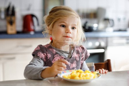 Foto de Hermosa niña comiendo papas fritas saludables para el almuerzo. Lindo bebé feliz niño en ropa colorida sentado en la cocina del hogar, guardería o guardería. El niño come verduras. - Imagen libre de derechos
