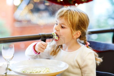 Téléchargez les photos : Petite fille d'âge préscolaire assise dans un restaurant près de la fenêtre décorée pour Noël. Un enfant qui mange des pâtes. Heureux temps en famille. - en image libre de droit
