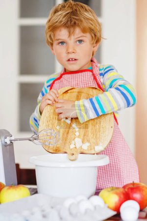 Foto de Lindo poco feliz niño rubio preescolar hornear pastel de manzana y magdalenas en la cocina doméstica. Divertido niño encantador saludable divertirse con el trabajo con mezclador, harina, huevos, frutas. Pequeño ayudante en interiores. - Imagen libre de derechos