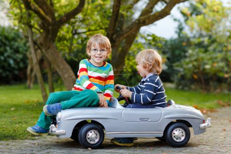 Téléchargez les photos : Deux petits garçons d'âge préscolaire jouant avec une grande vieille voiture jouet dans le jardin d'été, à l'extérieur. Des enfants heureux jouent ensemble, conduisant une voiture. Activité de plein air pour les enfants. Frères et amis par temps chaud. - en image libre de droit