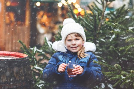 Téléchargez les photos : Petite fille préscolaire mignonne qui boit du punch chaud pour enfants ou du chocolat sur le marché de Noël allemand. Enfant heureux sur le marché familial traditionnel en Allemagne, Rire garçon en vêtements d'hiver colorés. - en image libre de droit