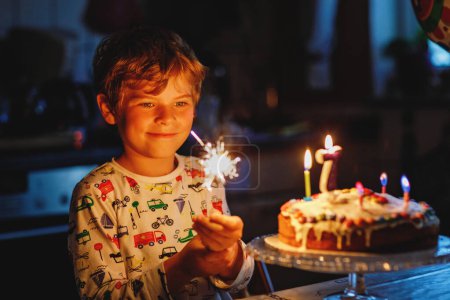 Téléchargez les photos : Adorable petit garçon blond joyeux célébrant son anniversaire. Enfant soufflant sept bougies sur un gâteau fait maison, à l'intérieur. Fête d'anniversaire pour les écoliers, célébration familiale de 7 ans. - en image libre de droit