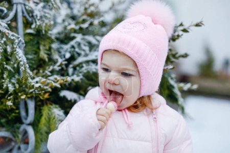 Photo for Adorable little toddler girl walking outdoors in winter. Cute toddler eating sweet lollypop candy. Child having fun on cold snow day. Wearing warm baby pink clothes and hat with bobbles - Royalty Free Image