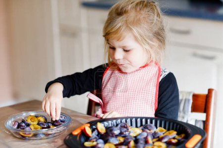 Téléchargez les photos : Mignon petit tout-petit fille cuisson tarte aux prunes à la maison. Heureux enfant souriant aider et préparer des prunes pour le gâteau dans la cuisine domestique. Aliments maison sains - en image libre de droit