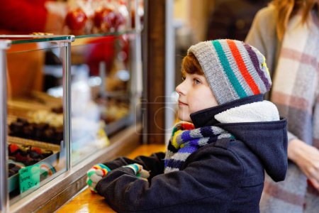 Téléchargez les photos : Petit garçon mignon près d'un stand sucré avec des pommes sucrées et des fruits au chocolat. Joyeux enfant sur le marché de Noël en Allemagne. Loisirs traditionnels pour les familles à Noël. Vacances, célébration, tradition - en image libre de droit