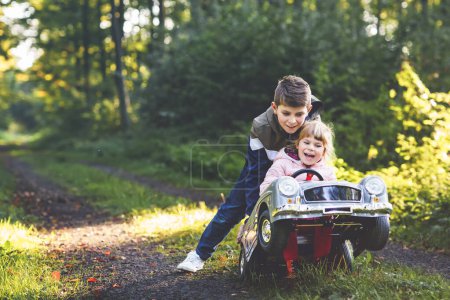 Foto de Dos niños felices jugando con un gran coche de juguete viejo en el bosque de otoño, al aire libre. Niño empujando y conduciendo coche con niña pequeña, hermana linda dentro. Riendo y sonriendo niños. Familia encantadora.. - Imagen libre de derechos