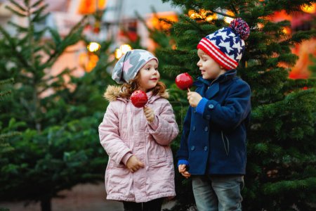 Foto de Dos niños sonrientes, niño y niña comiendo manzana azucarada cristalizada en el mercado navideño alemán. Amigos felices en ropa de invierno con luces en el fondo. Familia, tradición, concepto de vacaciones
. - Imagen libre de derechos