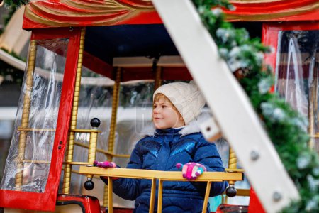 Téléchargez les photos : Joyeux mignon fille préscolaire chevauchant sur ferris roue carrousel cheval à Noël foire ou marché, en plein air. Petit enfant en bas âge s'amuser sur le marché de Noël familial traditionnel en Allemagne. - en image libre de droit