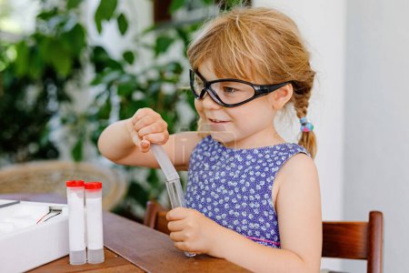 Foto de Una niña preescolar haciendo un experimento químico en el laboratorio de la escuela. Un niño pequeño con gafas protectoras estudia cómo dejar caer líquido a una sonda de ensayo, caucásica, bioquímica. clase de química - Imagen libre de derechos