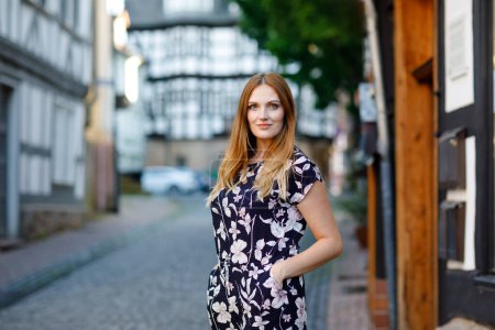 Téléchargez les photos : Belle jeune femme aux cheveux longs en robe d'été se promenant dans la ville allemande. Fille heureuse profitant de la marche dans une jolie petite ville fachwerk avec de vieilles maisons en Allemagne - en image libre de droit