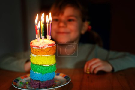 Foto de Happy little preschool girl celebrating birthday. Closeup of child with homemade rainbow cake, indoor. Happy healthy toddler blowing six candles on cake. Selective focus on cake. - Imagen libre de derechos