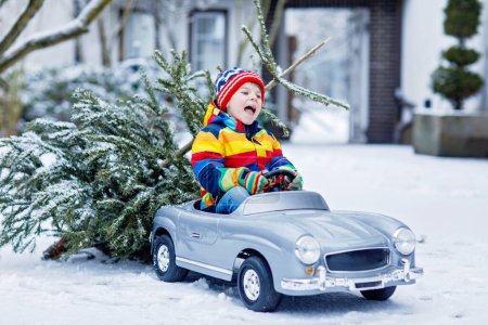 Téléchargez les photos : Petit garçon drôle et souriant conduisant une voiture jouet avec arbre de Noël. Enfant heureux en vêtements de mode d'hiver apportant arbre de Noël taillé de forêt enneigée. Famille, tradition, vacances - en image libre de droit