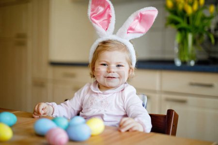 Foto de Linda niña pequeña con orejas de conejo de Pascua jugando con huevos pastel de colores. Niño feliz desempacando regalos. Adorable niño sonriente sano en ropa rosa disfrutando de unas vacaciones en familia, - Imagen libre de derechos