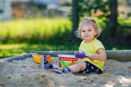 Foto de Feliz niña jugando en la arena en el patio al aire libre. Bebé divirtiéndose en el soleado verano cálido día soleado. Niño activo con juguetes de arena y en ropa de moda colorida - Imagen libre de derechos