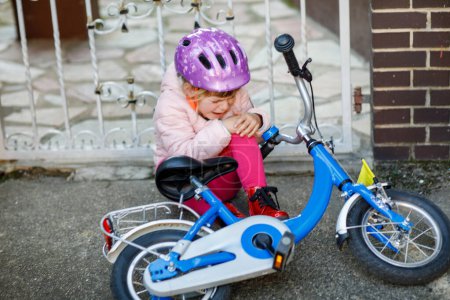 Photo for Cute little girl sitting on the ground after falling off her bike. Upset crying preschool child with safe helmet getting hurt while riding a bicycle. Active family leisure with kids - Royalty Free Image