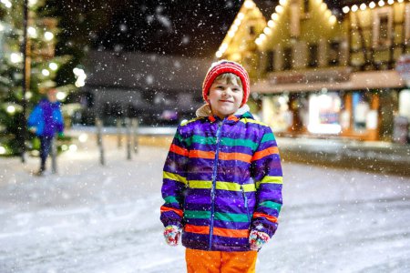 Téléchargez les photos : Petit garçon drôle en vêtements colorés jouant à l'extérieur pendant les chutes de neige. Loisirs actifs avec les enfants en hiver par temps froid et neigeux. Joyeux enfant sur le marché de Noël en Allemagne - en image libre de droit