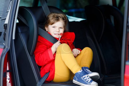 Photo for Little toddler girl sitting in car seat, holding sweet lollipop against nausea and sickness and looking out of the window on nature and traffic. Cute kid traveling by car. Child safety on the road - Royalty Free Image
