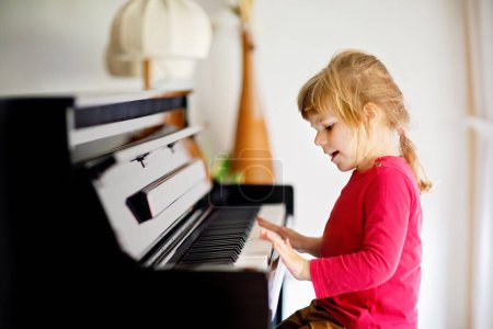 Foto de Hermosa niña tocando el piano en la sala de estar. Lindo niño preescolar que se divierte con aprender a tocar el instrumento de música. Educación musical temprana para niños - Imagen libre de derechos