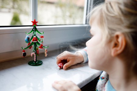 Foto de Niña pequeña sentada junto a la ventana y decorando un pequeño árbol de Navidad de vidrio con pequeños juguetes de Navidad. Feliz niño sano celebrar la fiesta tradicional familiar. Adorable bebé. - Imagen libre de derechos