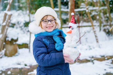 Photo for Cute little preschool girl with glasses making mini snowman. Adorable healthy happy child playing and having fun with snow, outdoors on cold day. Active leisure with children in winter. - Royalty Free Image