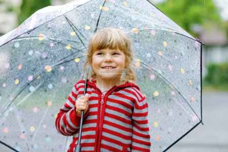 Foto de Pequeña niña jugando con un gran paraguas en el día lluvioso. Feliz niño positivo corriendo a través de la lluvia, charcos. Niño preescolar con ropa de lluvia y botas de goma. Actividad de los niños en el mal tiempo - Imagen libre de derechos