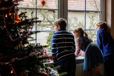 Foto de Tres lindos niños sentados junto a la ventana en Nochebuena. Dos niños de la escuela y una niña pequeña, hermanos mirando al aire libre y soñando. Felicidad familiar en vacaciones tradicionales. - Imagen libre de derechos