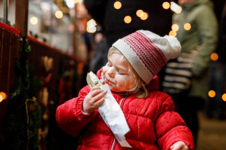 Téléchargez les photos : Petite fille, mignon enfant mangeant des bananes recouvertes de chocolat, des marshmellows et des saupoudres colorées près de stand sucré avec du pain d'épice et des noix. Joyeux bambin sur le marché de Noël en Allemagne - en image libre de droit