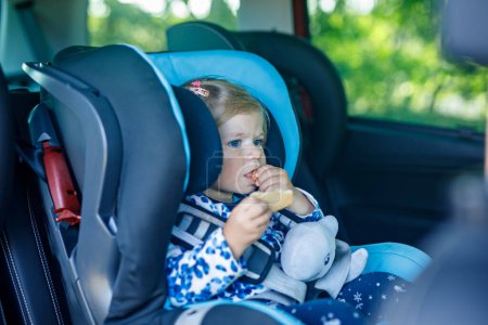 Téléchargez les photos : Adorable petite fille aux yeux bleus assise dans le siège de sécurité de la voiture. Enfant en bas âge en vacances en famille et Jorney. Sourire enfant heureux pendant les embouteillages, manger bisquit. - en image libre de droit