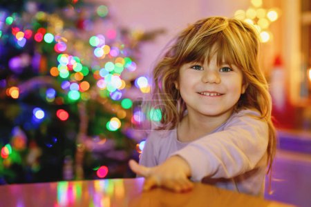 Foto de Niña pequeña sentada en pijama en la mañana de Navidad o víspera y con árbol de Navidad y luces en el fondo. Feliz niño sano celebrar la fiesta tradicional de la familia, esperando regalos - Imagen libre de derechos